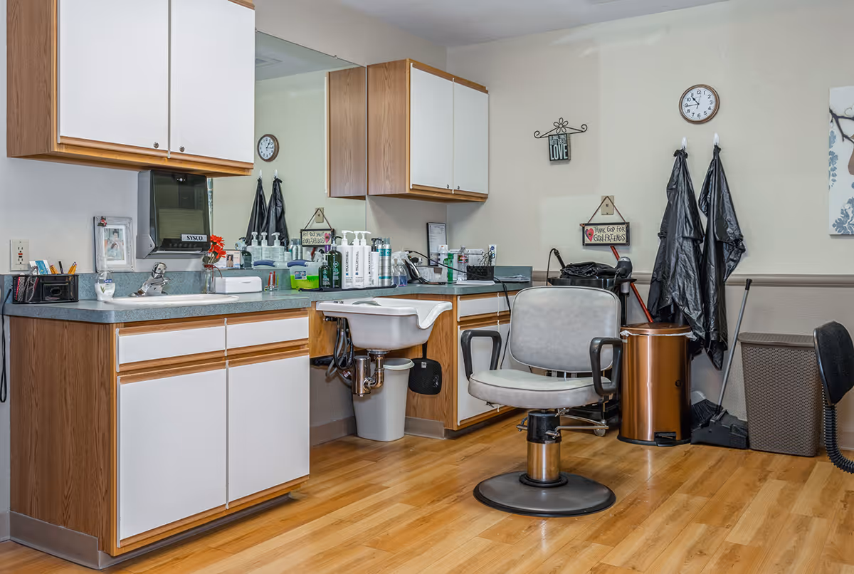 Interior view of a senior living facility's grooming or salon area featuring a salon chair in front of a sink, wooden cabinets with white doors, a large mirror, various bottles and grooming supplies on the countertop, a clock on the wall, and some hanging black aprons.