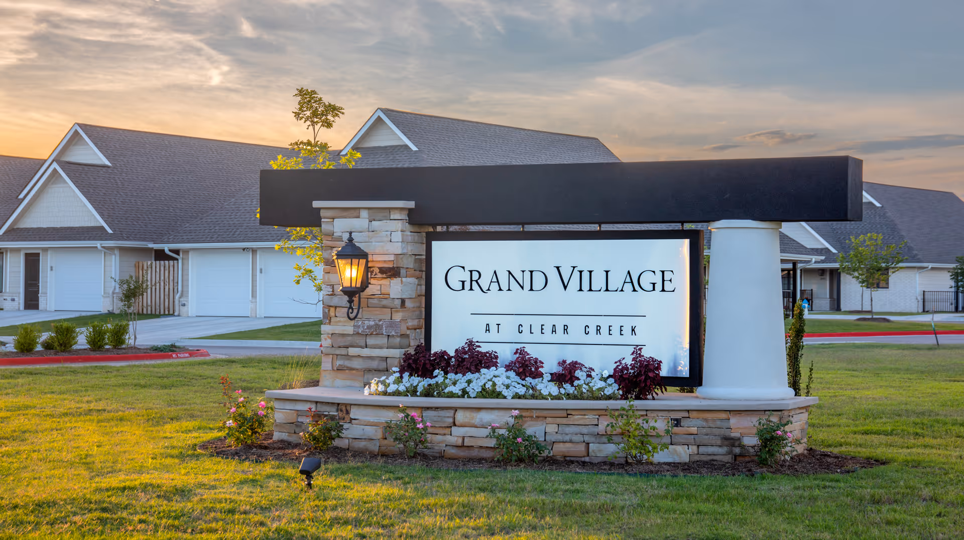 Entrance sign for Grand Village at Clear Creek with a stone and white pillar base, surrounded by flowers and grass, with residential buildings in the background under a partly cloudy sky at sunset.