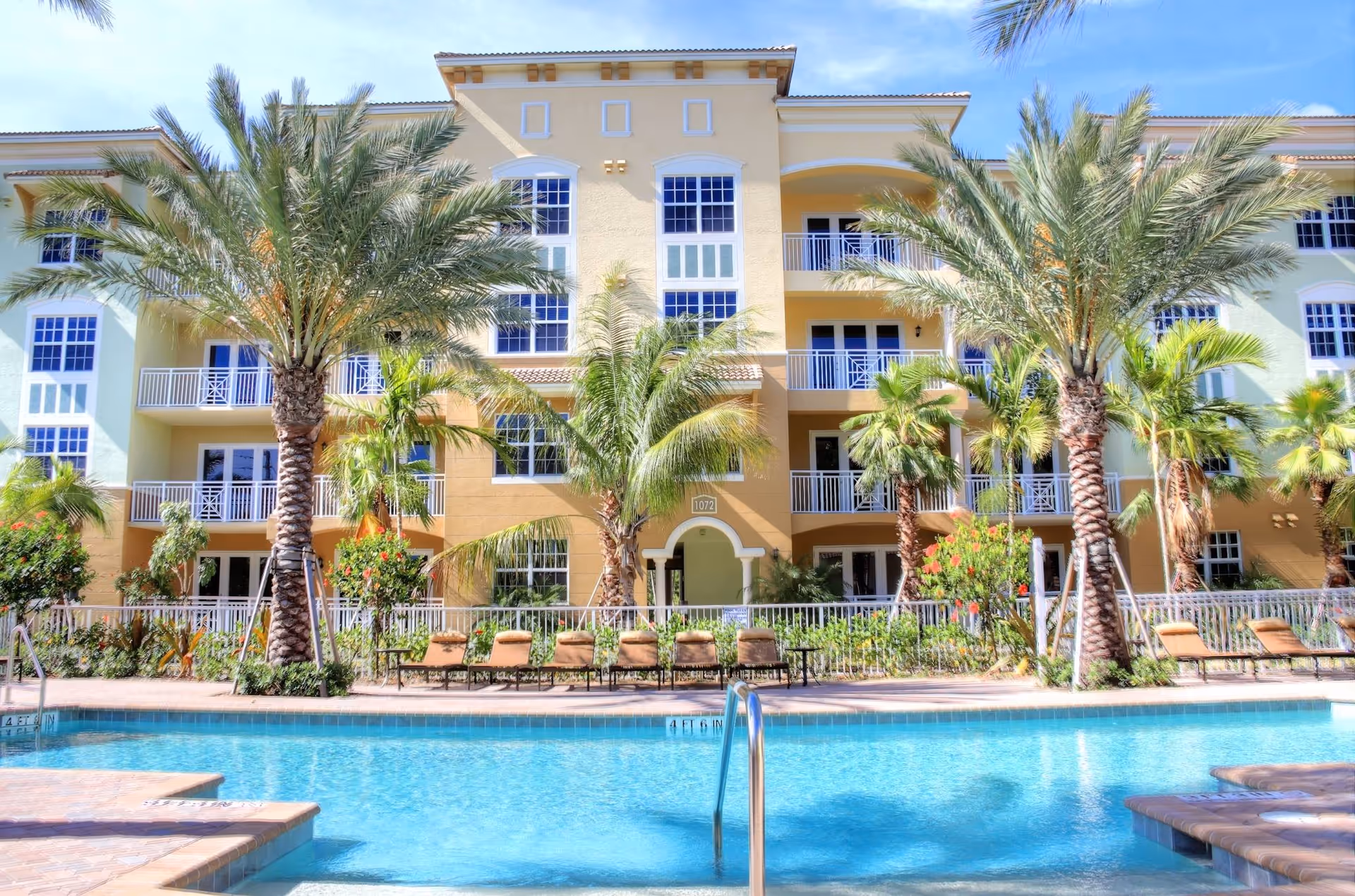 Outdoor swimming pool and lounge area in front of a multi-story Mediterranean-style building with balconies and palm trees.