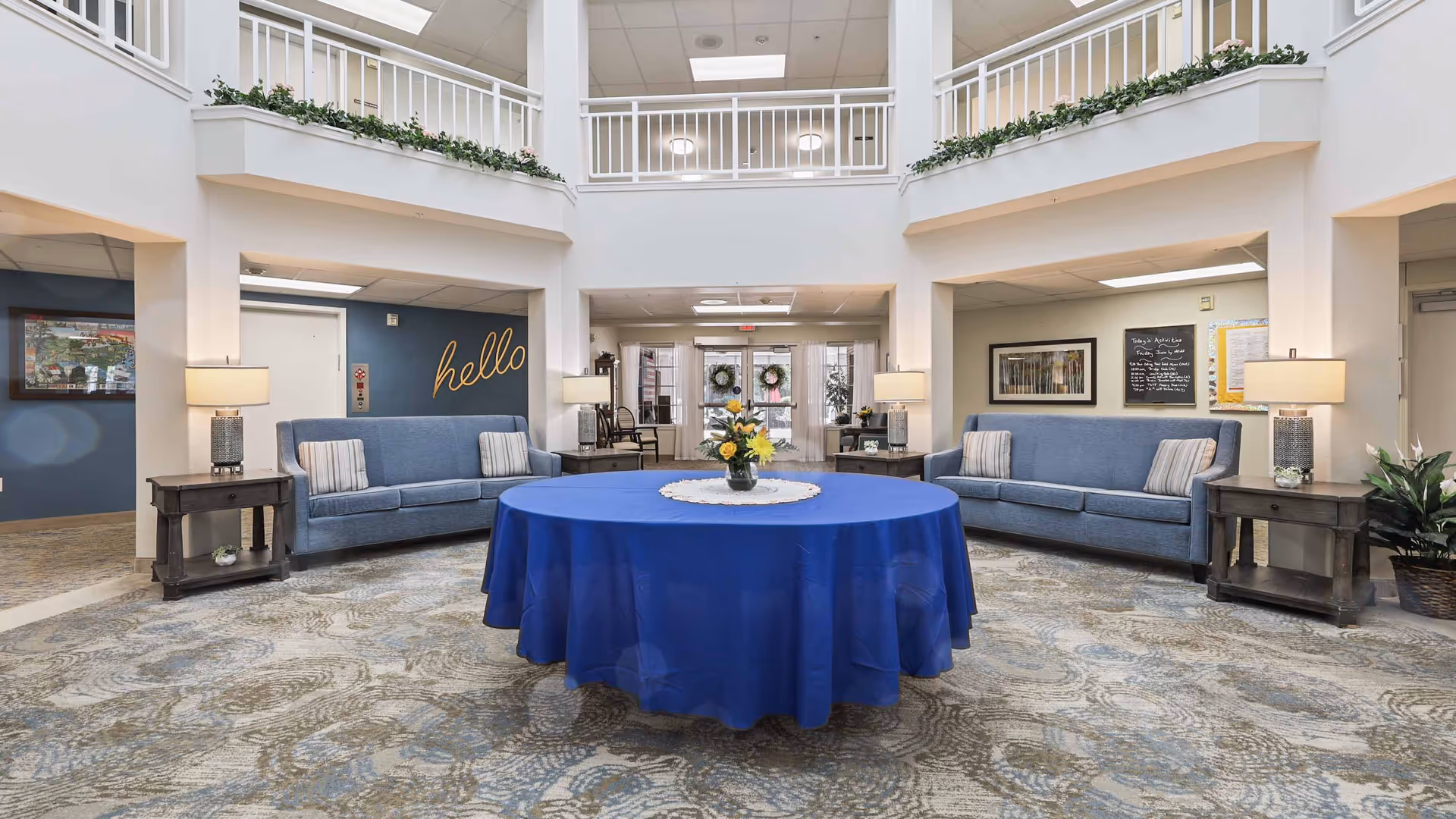 A spacious and well-lit common area in a senior living facility with two blue sofas facing each other, each with two striped pillows. A round table covered with a blue tablecloth and a flower arrangement is centered on a patterned carpet. The walls are white with a second-floor balcony railing visible above. There are lamps on dark wooden side tables next to each sofa, and a wall with the word 'hello' written in cursive is visible in the background.