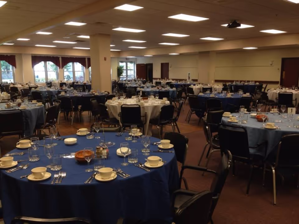 Large dining hall with round tables set for a meal, covered in blue and white tablecloths and place settings.