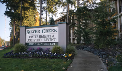 Outdoor view of the entrance sign for Silver Creek Retirement & Assisted Living located at 17607 91st Avenue East, surrounded by greenery and trees with a building partially visible in the background.