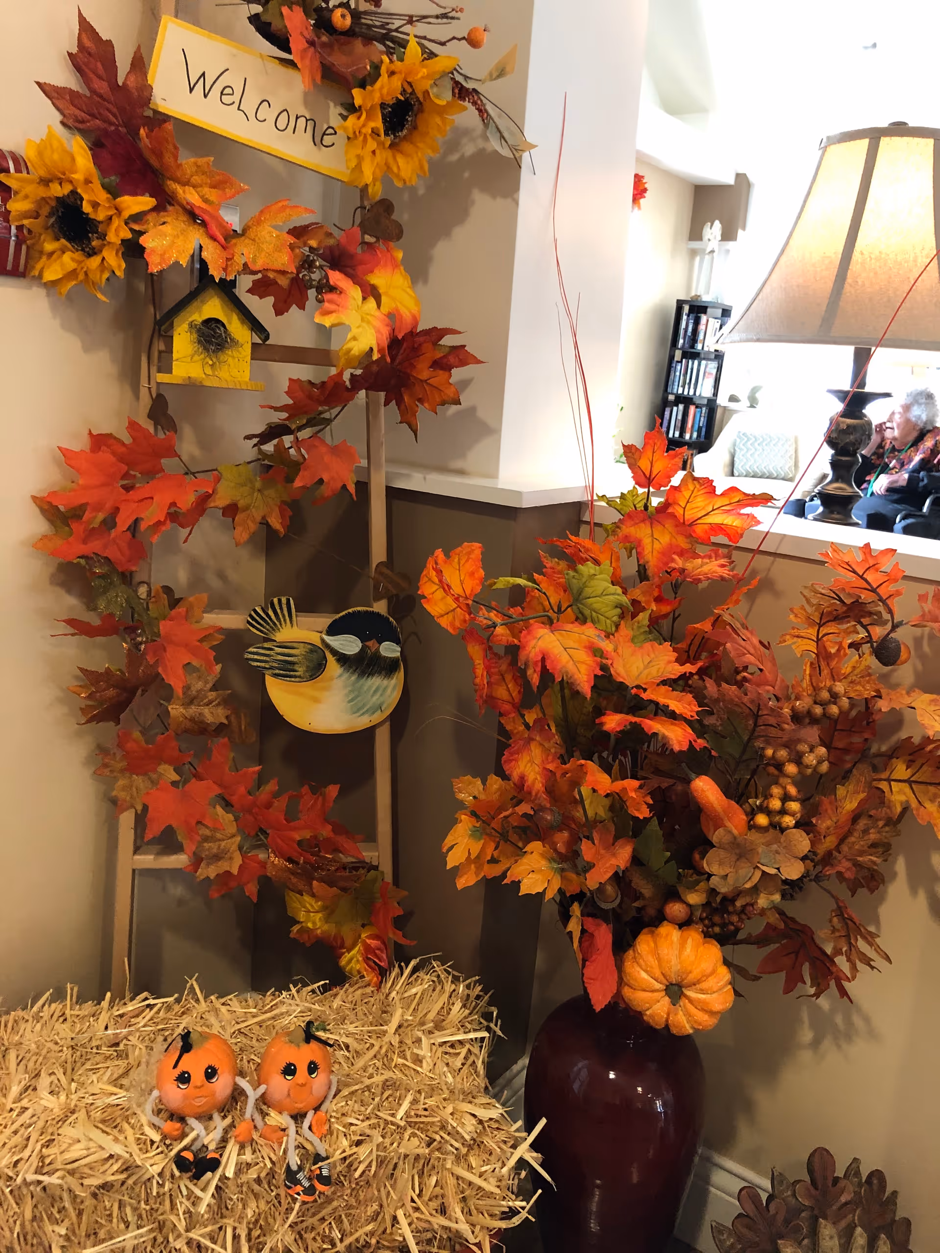 A cozy interior corner decorated for fall with a vase of orange leaves, garland, small pumpkin dolls on a straw bale, and a lamp-lit seating area visible in the background.