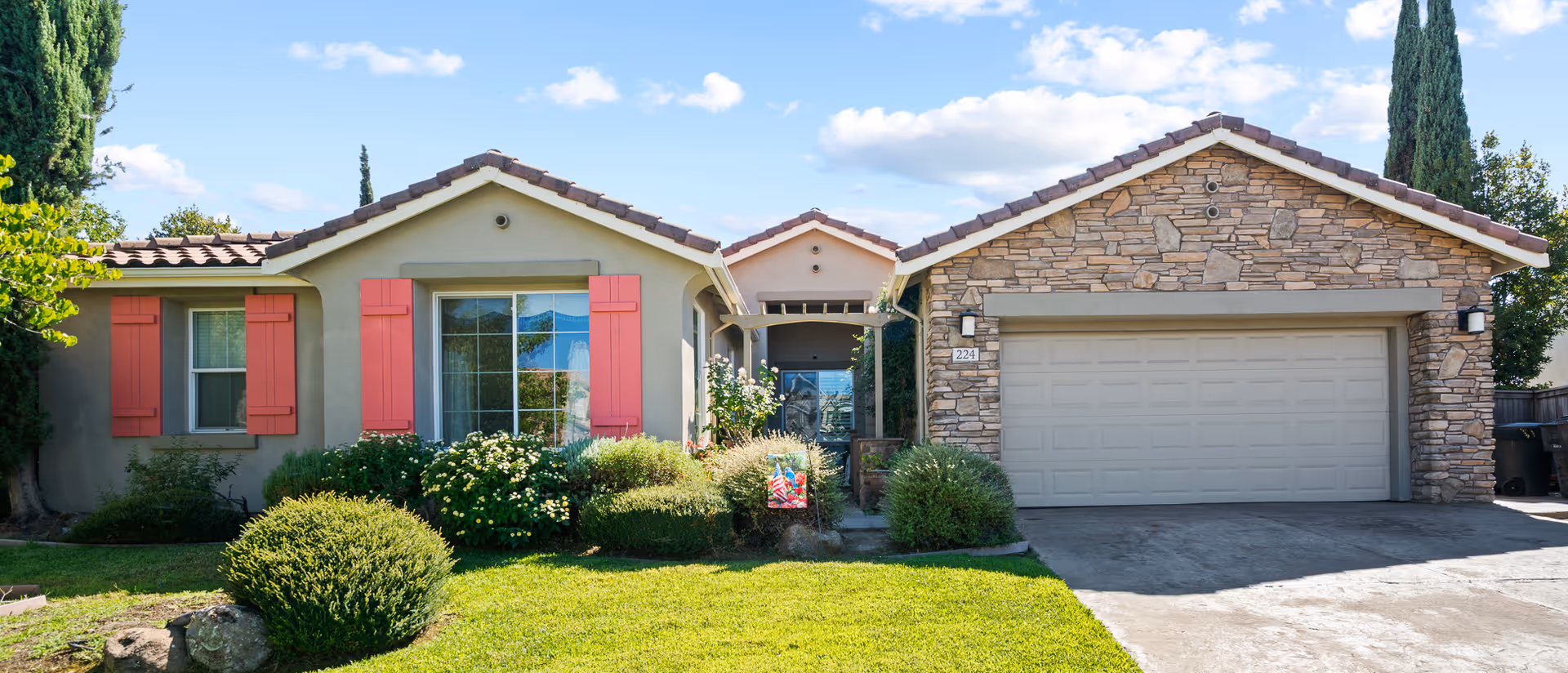 Front exterior view of a single-story house with beige walls, red window shutters, a stone facade garage, and a well-maintained green lawn with bushes and trees under a blue sky with some clouds.
