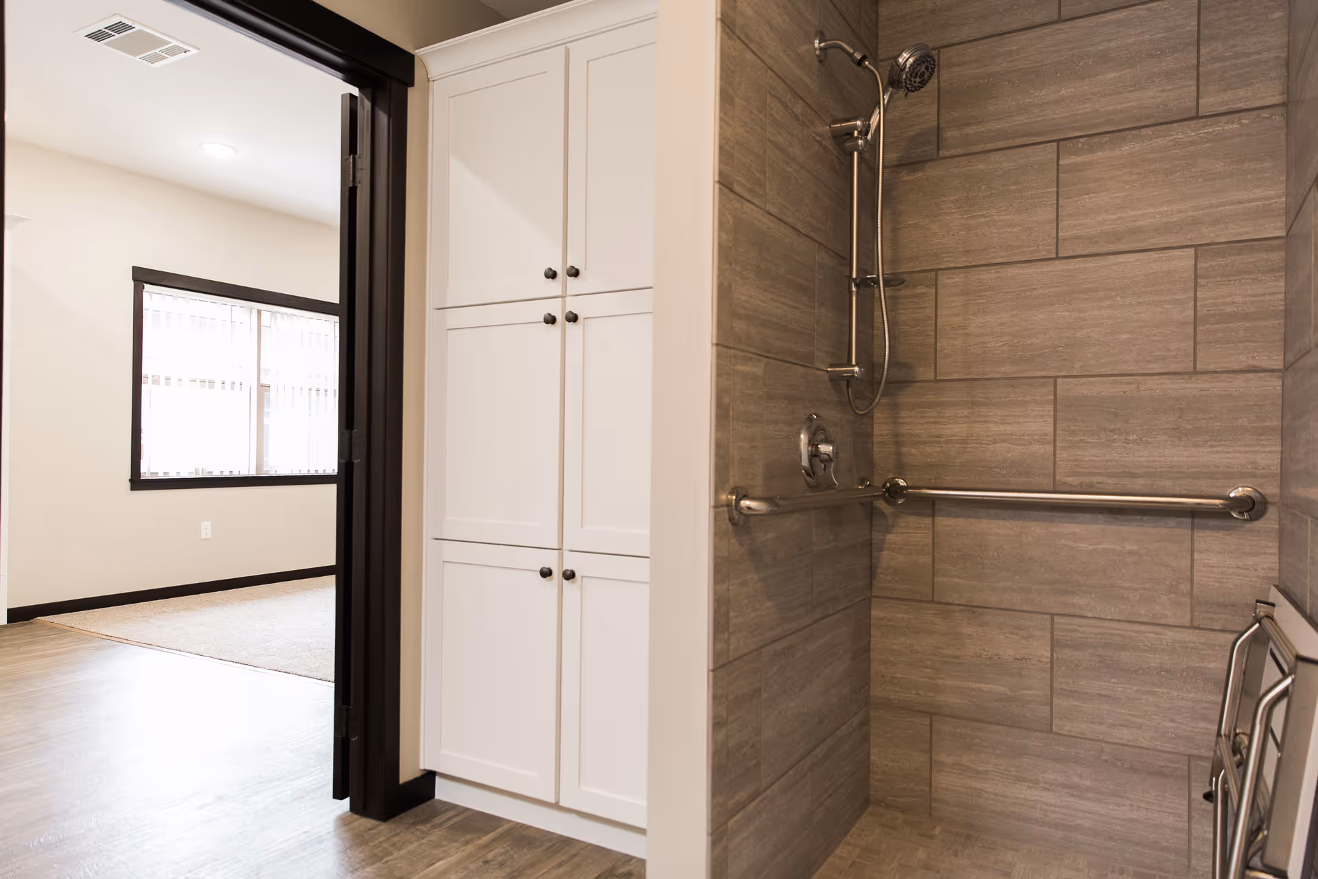 A modern bathroom shower area with gray tiled walls and a stainless steel grab bar. Adjacent to the shower is a white cabinet with four doors. The image also shows a doorway leading to a bright room with a large window and light-colored walls.