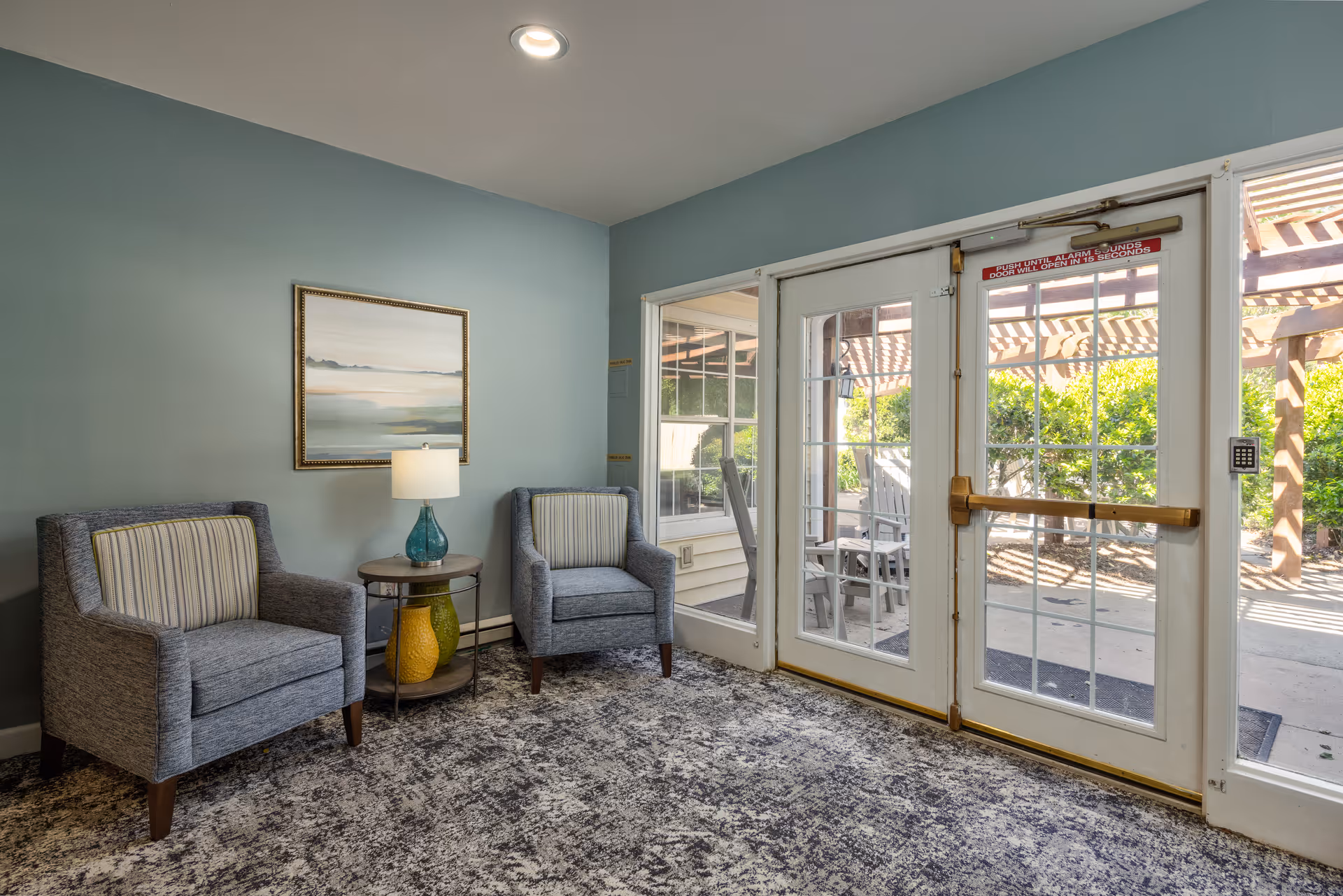 A small seating area with two gray armchairs with striped cushions, a round side table with a blue lamp and decorative vases, a framed landscape painting on a light blue wall, and glass double doors leading to an outdoor patio with wooden pergola and greenery.