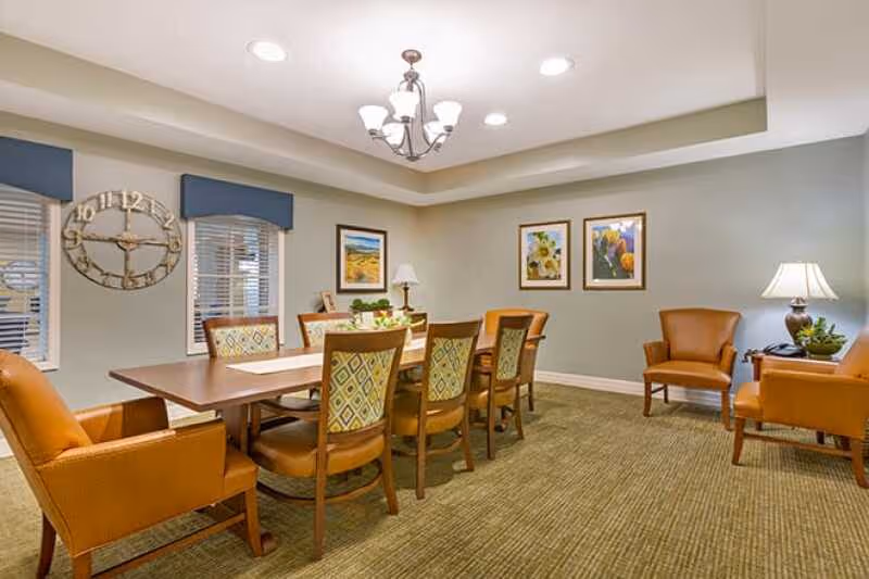 Well-lit communal dining room with a long wooden table surrounded by upholstered chairs, artwork on the walls, and a decorative clock.