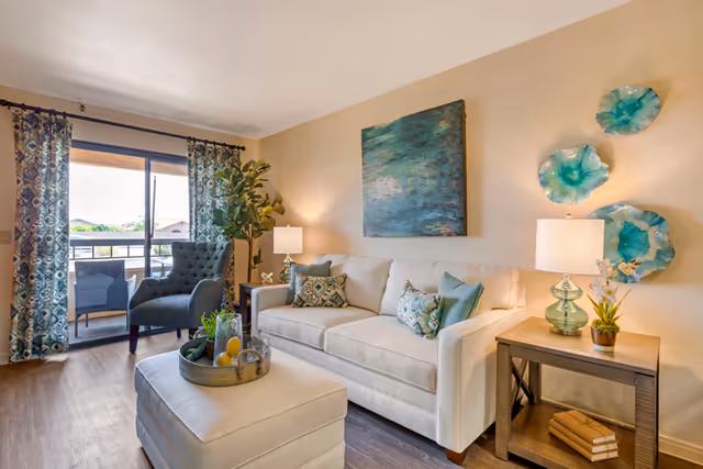 A bright living room with a white sofa adorned with patterned and solid teal pillows, a matching ottoman with a decorative tray, a side table with a lamp and small plant, and three teal decorative wall hangings. A gray armchair is positioned near a sliding glass door with patterned curtains leading to a balcony with outdoor seating and a view of neighboring houses.