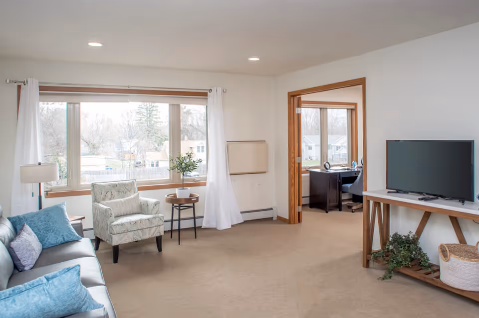 Bright living room with a sofa, armchair, large window, TV on a console and a doorway leading to an office.