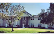 Exterior view of a two-story assisted living facility building with white siding and a central stone facade entrance, surrounded by green grass and trees under a clear blue sky.