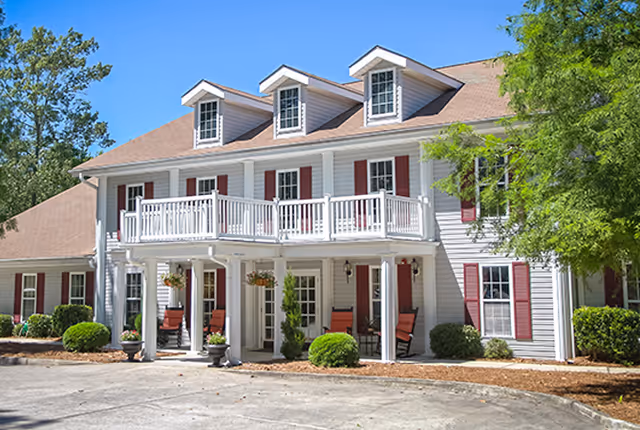 Exterior view of a two-story senior living facility building with white siding, red shutters, and a brown roof. The building features a balcony with white railings on the second floor and a covered porch area with seating on the ground floor. There are green shrubs and trees around the building under a clear blue sky.