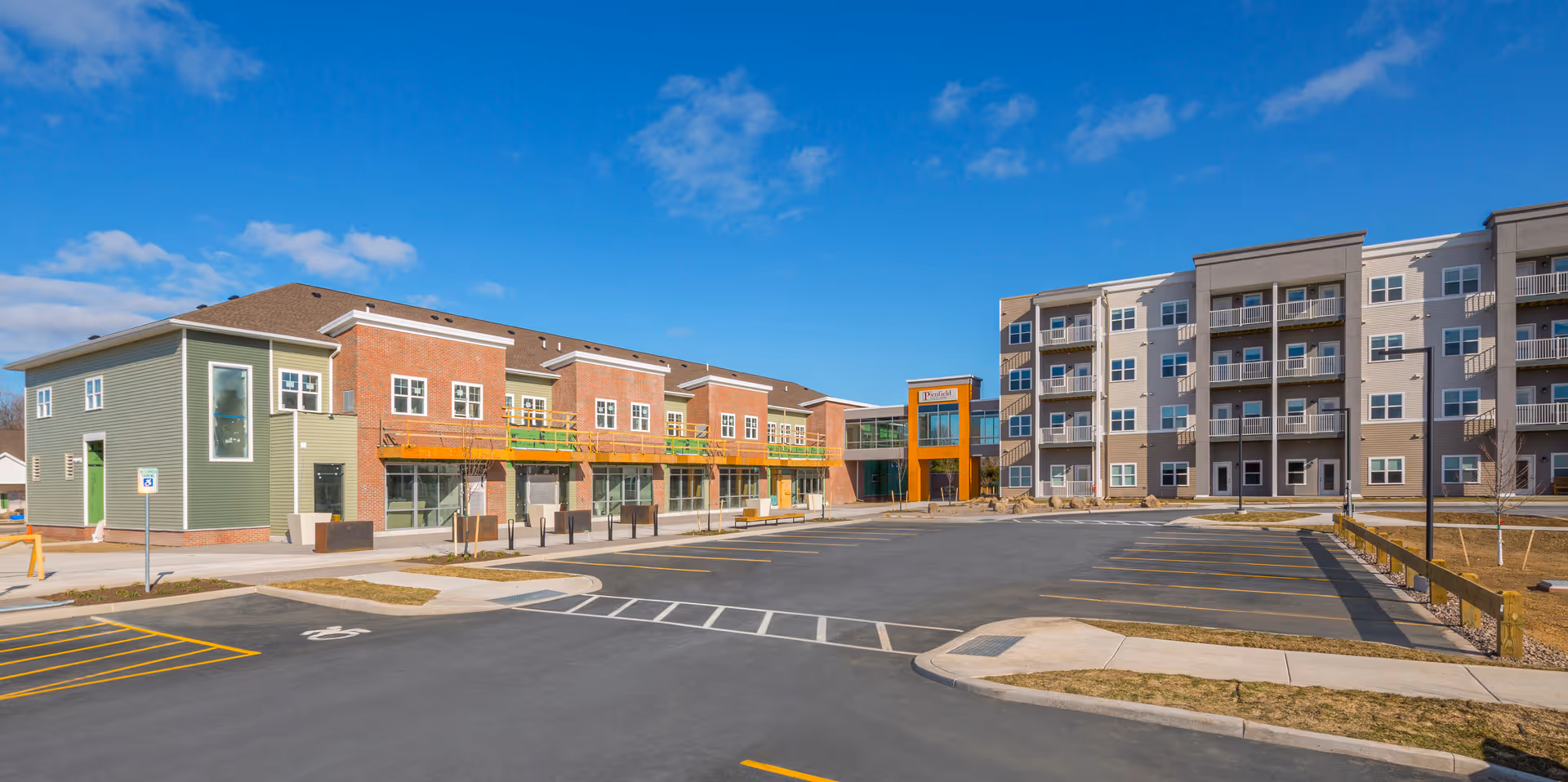 Exterior view of a senior living facility with a large parking lot in front. The building has multiple sections with different architectural styles, including a green and brick section with balconies and a taller beige section with multiple balconies. The sky is clear with a few clouds.