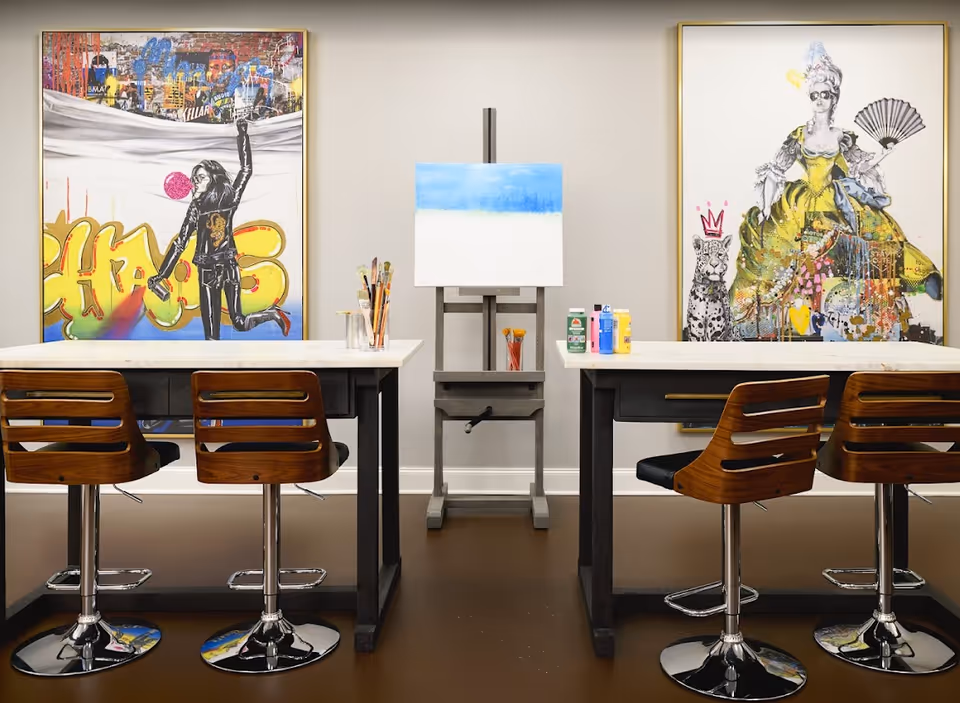Bright art studio/activity room with two high tables, four wooden swivel stools, an easel with a canvas, paint supplies, and colorful framed artwork on the wall.