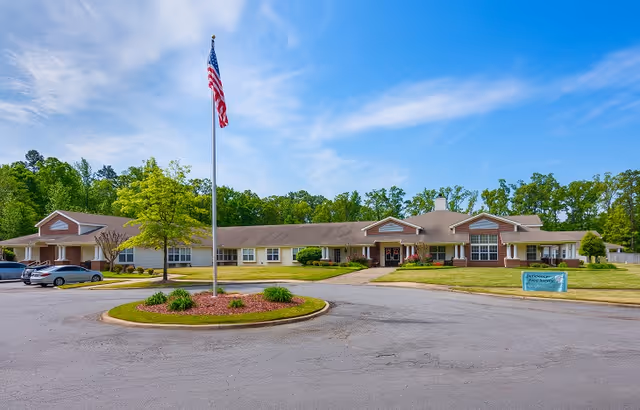 Single-story senior living facility building with a large parking area in front, an American flag on a flagpole in the center of a circular landscaped island, and a sign on the right side of the lawn. The building is surrounded by trees under a partly cloudy blue sky.