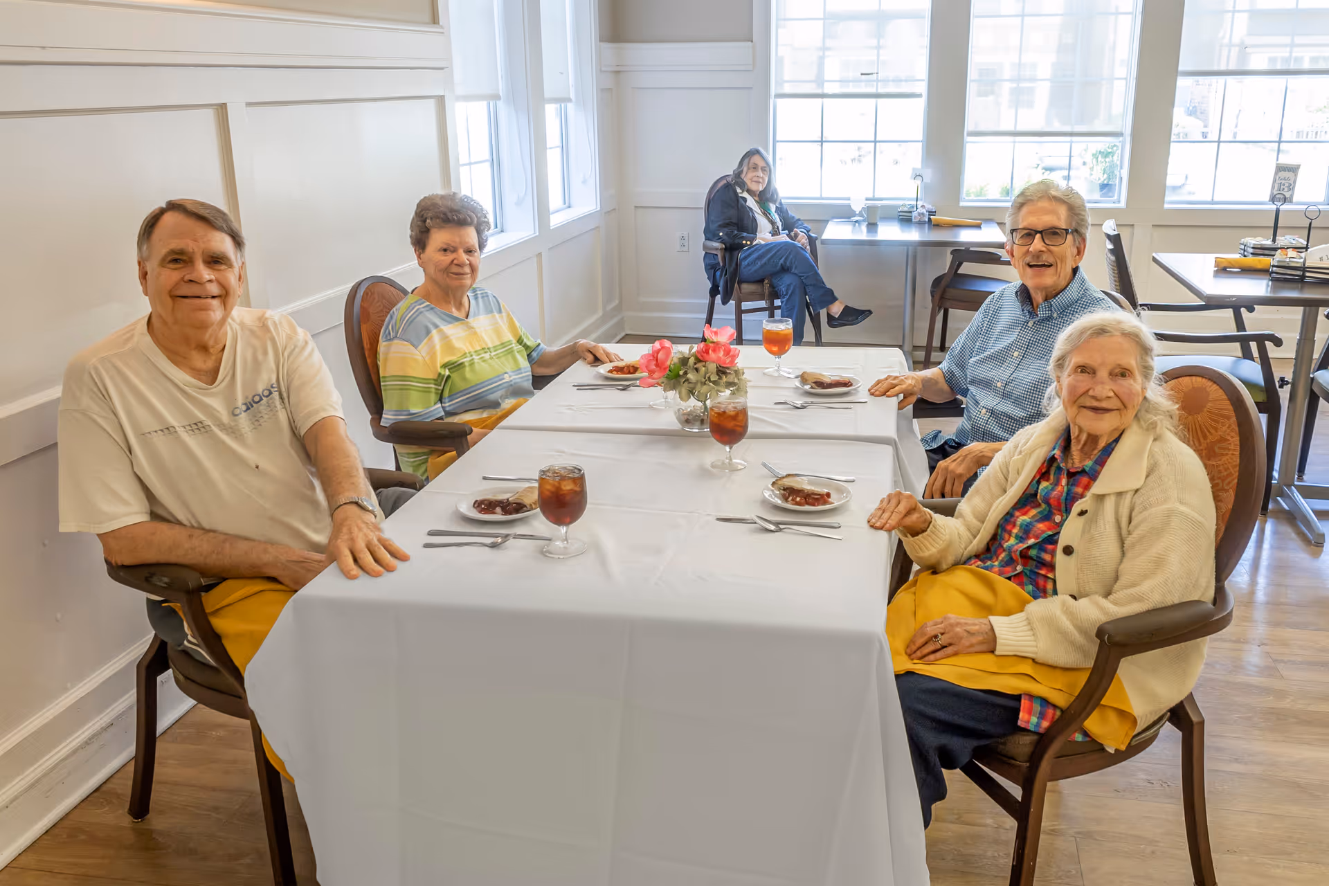 Five older adults sit around a dining table set with iced tea and plates in a bright dining room.
