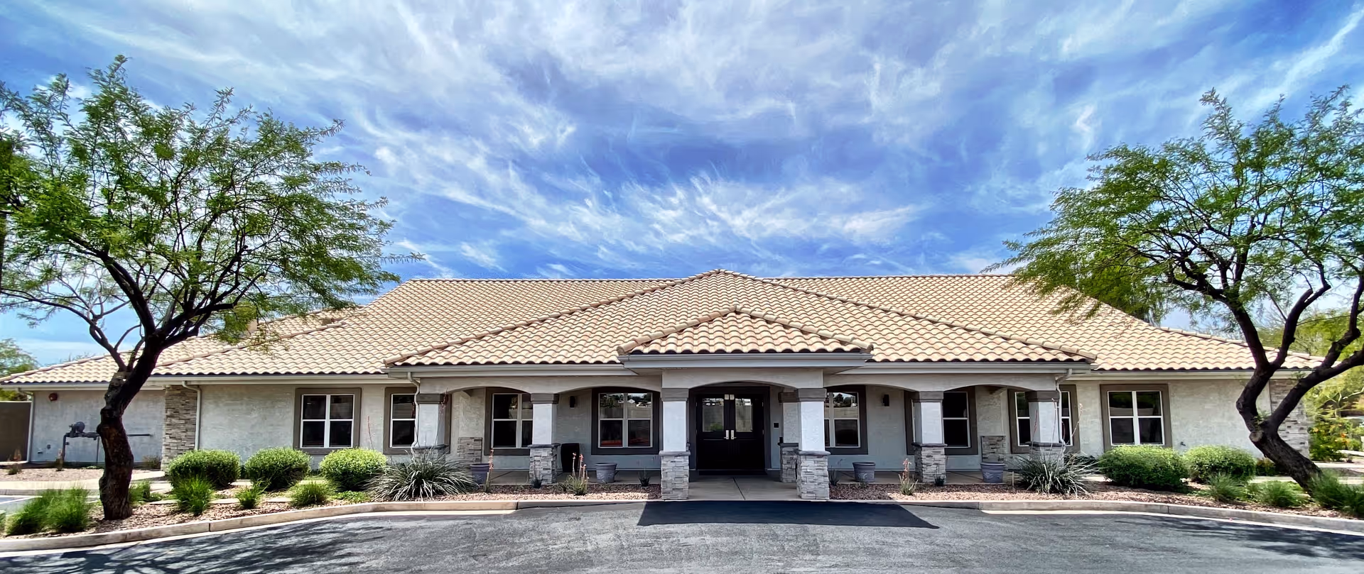 Front exterior view of a single-story building with a tiled roof, multiple windows, and a covered entrance supported by stone pillars. The building is surrounded by landscaped greenery including bushes and two trees, under a partly cloudy blue sky.