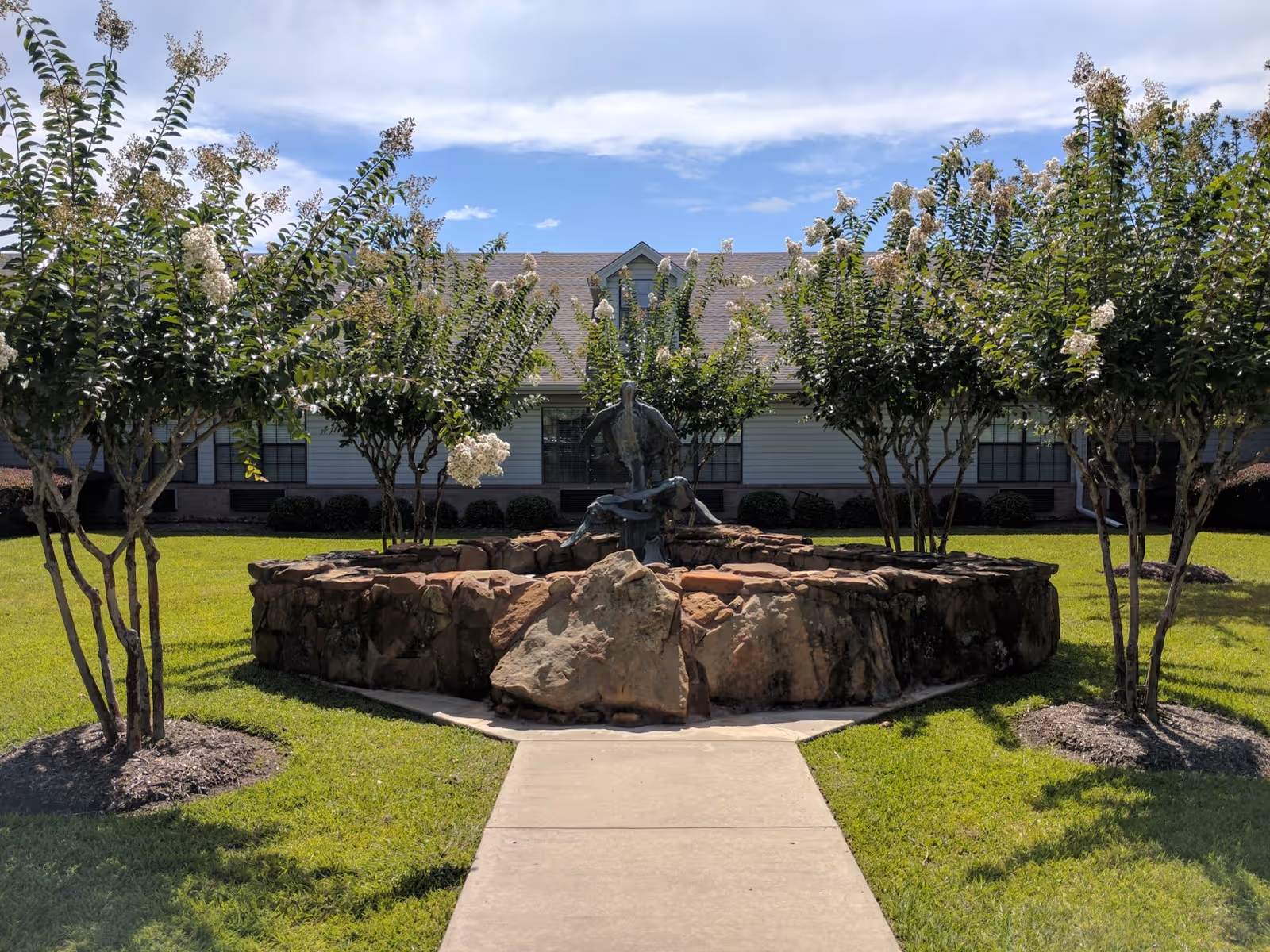 A sidewalk leads to a circular stone fountain with a bronze sculpture, surrounded by flowering trees and a one-story building in the background.