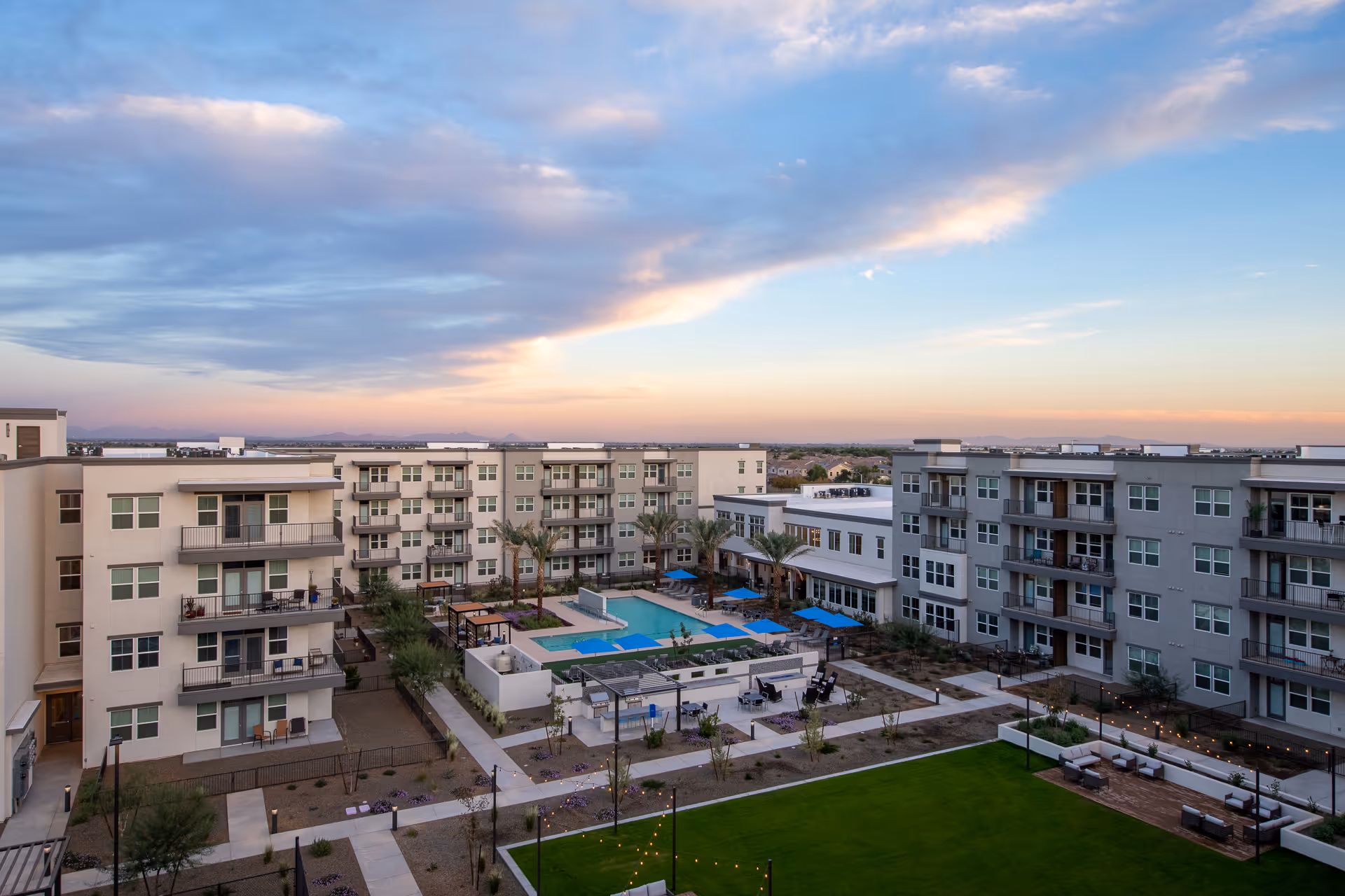 Aerial view of a modern residential complex at sunset featuring multiple apartment buildings surrounding a central courtyard with a swimming pool, blue umbrellas, palm trees, walkways, and outdoor seating areas with string lights.