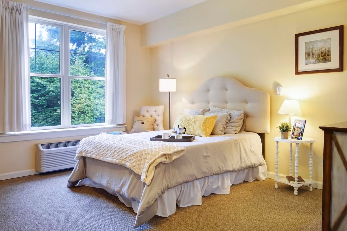 A cozy bedroom with a large window showing green trees outside. The room features a bed with a tufted headboard, multiple pillows, a knitted blanket, and a tray with a coffee cup. There is a cushioned chair in the corner, a floor lamp, a small round side table with a lamp, a framed photo, and a small plant. A framed picture hangs on the wall above the side table.