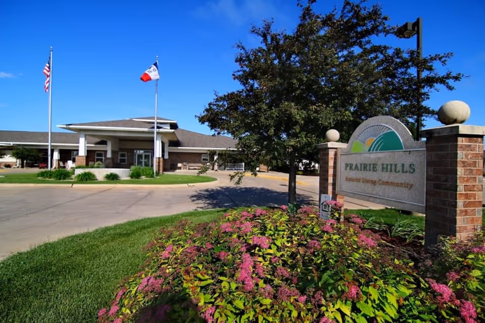 Front entrance of Prairie Hills assisted living community with flags, landscaped flowerbeds, and the facility sign next to the driveway.