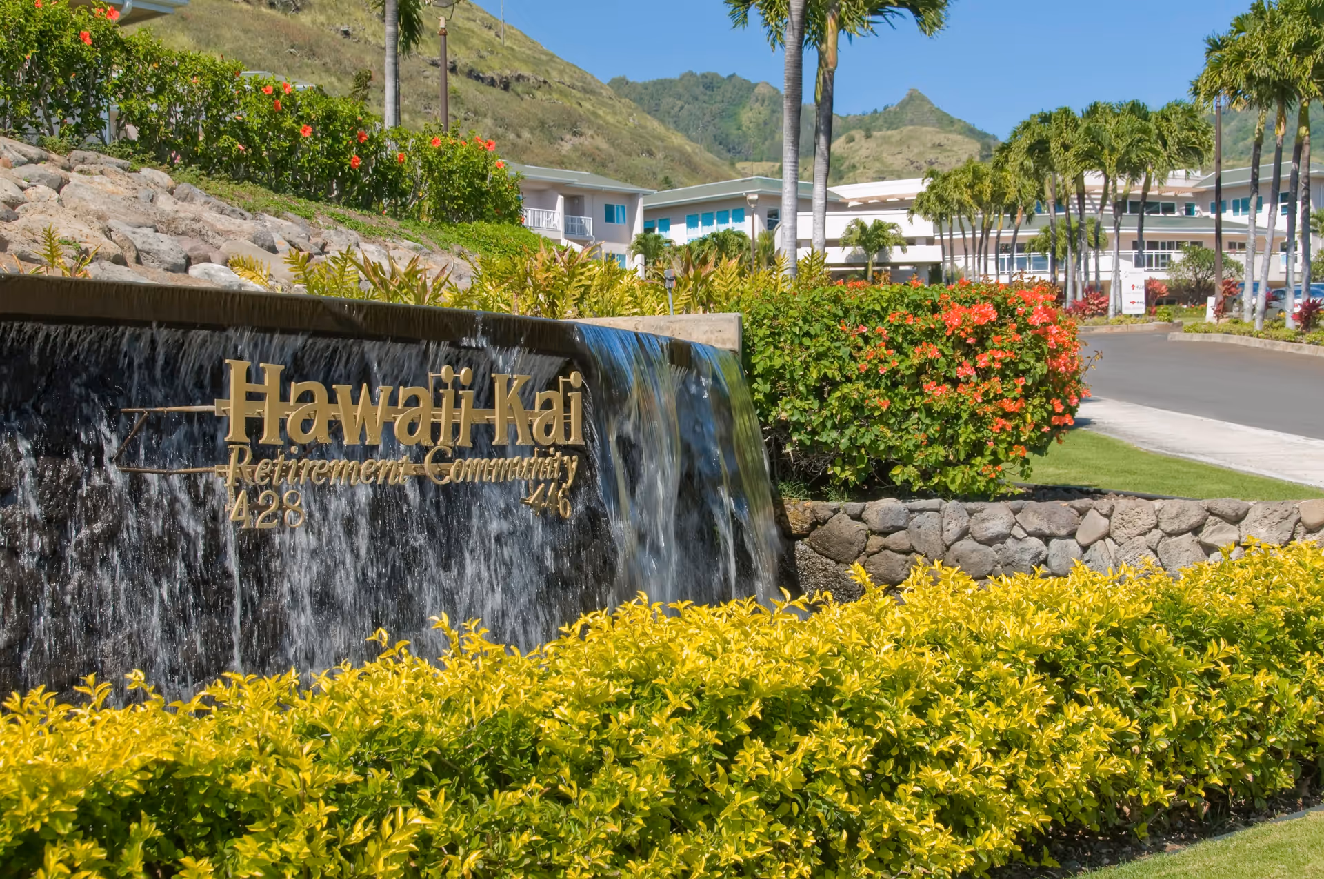 Outdoor view of the entrance to Hawaii Kai Retirement Community featuring a cascading water wall with the community's name in gold letters, surrounded by lush green and flowering plants, palm trees, and a clear blue sky with hills in the background.