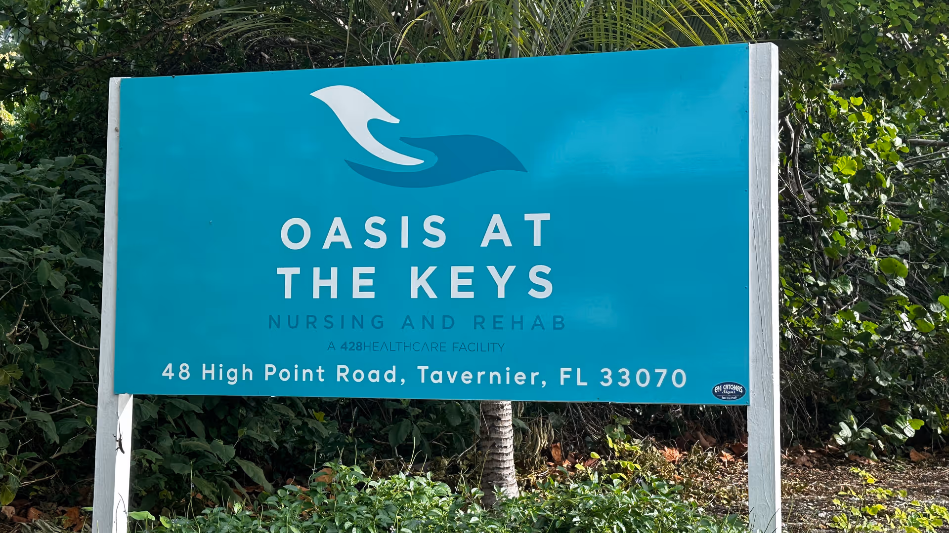 A teal-colored outdoor sign for Oasis at the Keys Nursing and Rehab facility, located at 48 High Point Road, Tavernier, FL 33070, surrounded by greenery and trees.