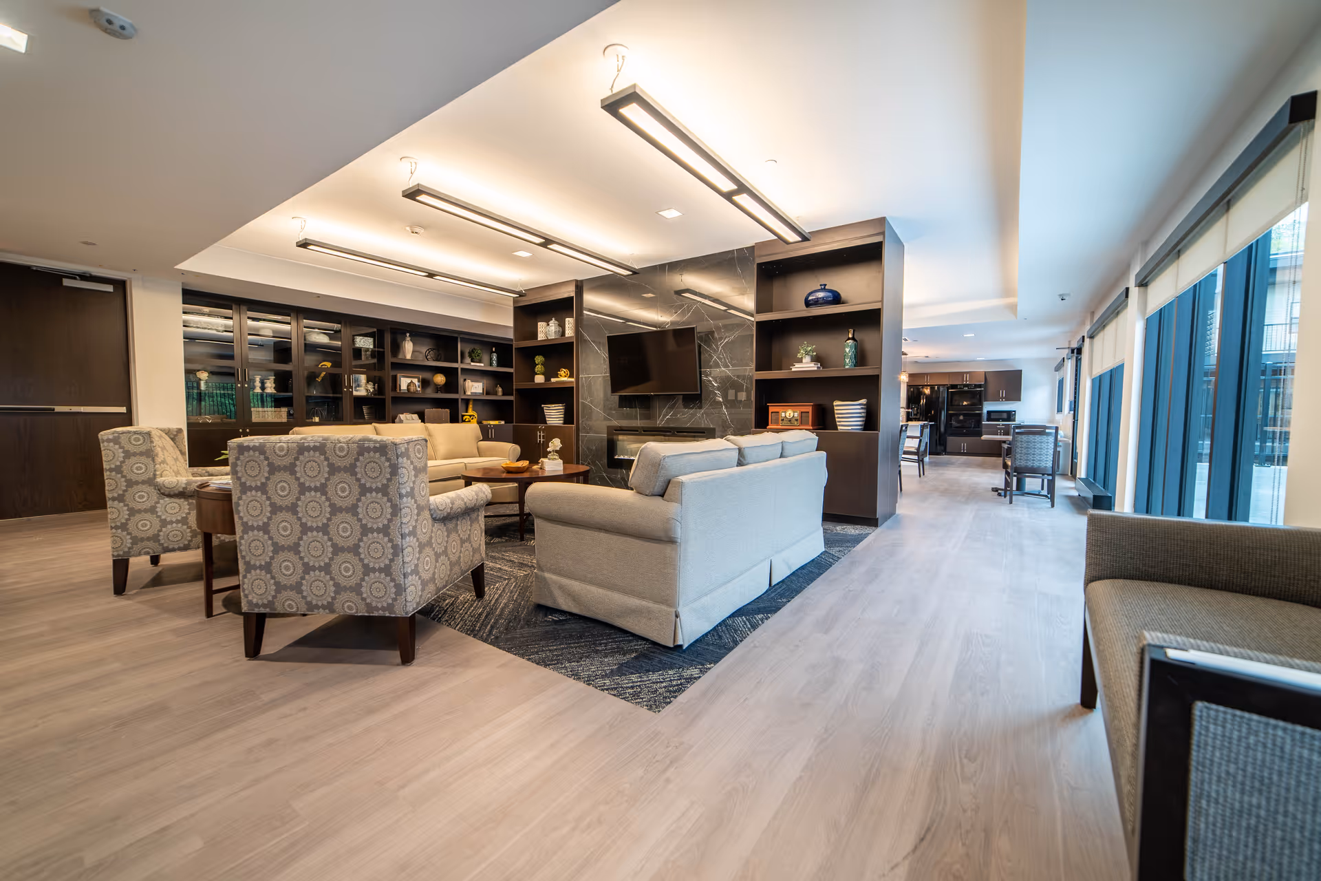 A spacious and modern living room area in a senior living facility featuring comfortable beige sofas and patterned armchairs arranged around a wooden coffee table. The room has built-in dark wood shelves with decorative items and a wall-mounted flat-screen TV above a sleek black fireplace. Large windows with roller shades line one side of the room, allowing natural light to fill the space. The flooring is light wood, and the ceiling has contemporary rectangular light fixtures.