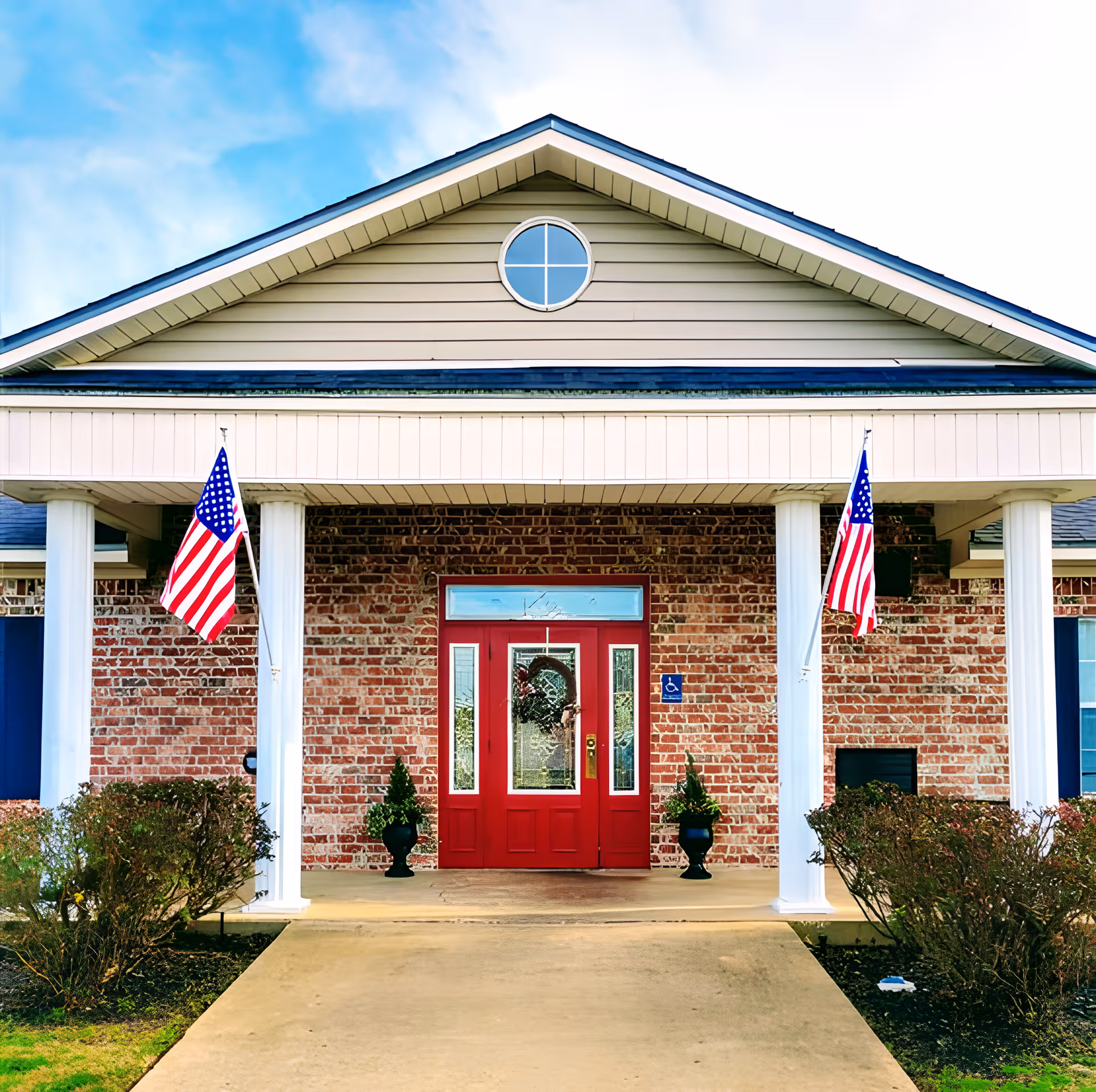 Front entrance of a brick building with a red double door, two white columns supporting a porch roof, two American flags on either side, and small bushes lining the walkway.