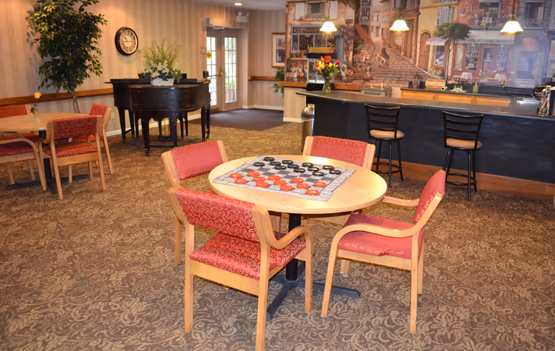 A senior living common dining/lounge area with a round table set for checkers, red cushioned chairs, a piano, and a bar counter with stools.