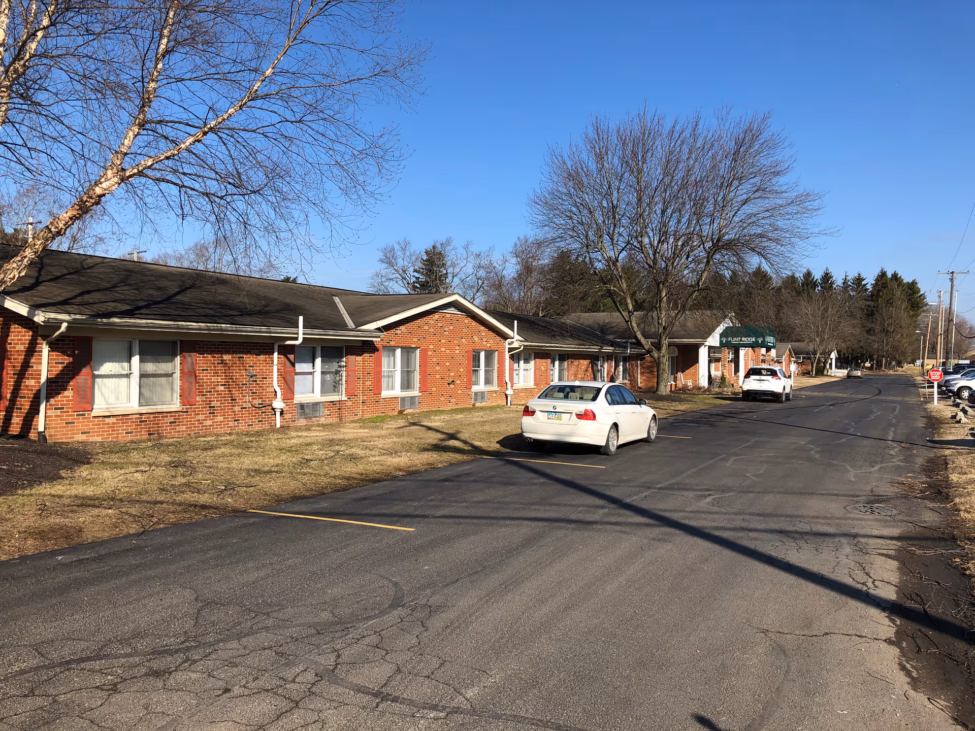 Exterior view of a single-story brick building with multiple windows and a black shingled roof, identified as Flint Ridge Nursing and Rehabilitation. There are two cars parked on the paved driveway in front of the building, leafless trees, and a clear blue sky.