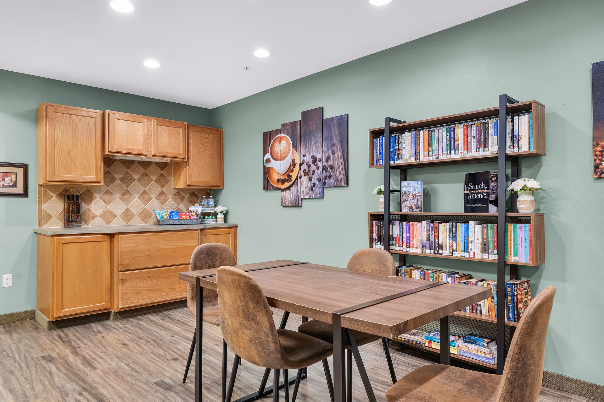 A cozy room with a wooden table and four brown chairs. On the right side, there is a tall bookshelf filled with books and some small potted plants. The back wall is painted green and decorated with a multi-panel artwork of a coffee cup and coffee beans. On the left side, there is a wooden cabinet with a tiled backsplash and snacks placed on the countertop.