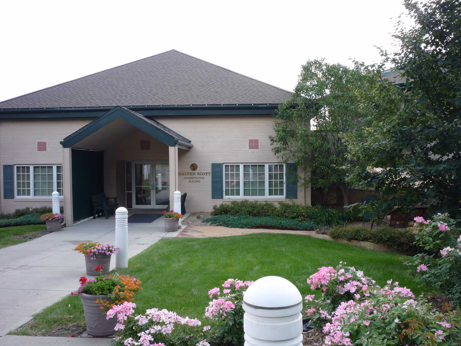 Exterior view of a single-story building with a peaked roof and a covered entrance. The building has beige walls with green window shutters and is surrounded by a well-maintained garden with green grass, flowering plants, and trees. There are potted flowers along the walkway leading to the entrance.