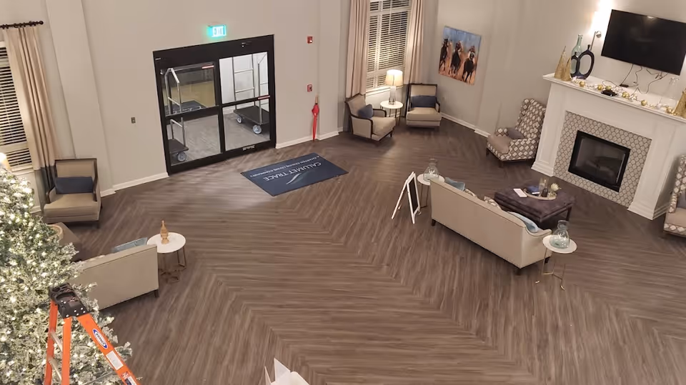 A spacious senior living community lobby with wood-patterned flooring, several seating areas including armchairs and sofas, a fireplace with a mounted TV above it, and a decorated Christmas tree with a ladder nearby. There is a glass exit door with an illuminated exit sign above it and a welcome mat that reads 'Calumet Trace - A Charter Senior Living Community'.