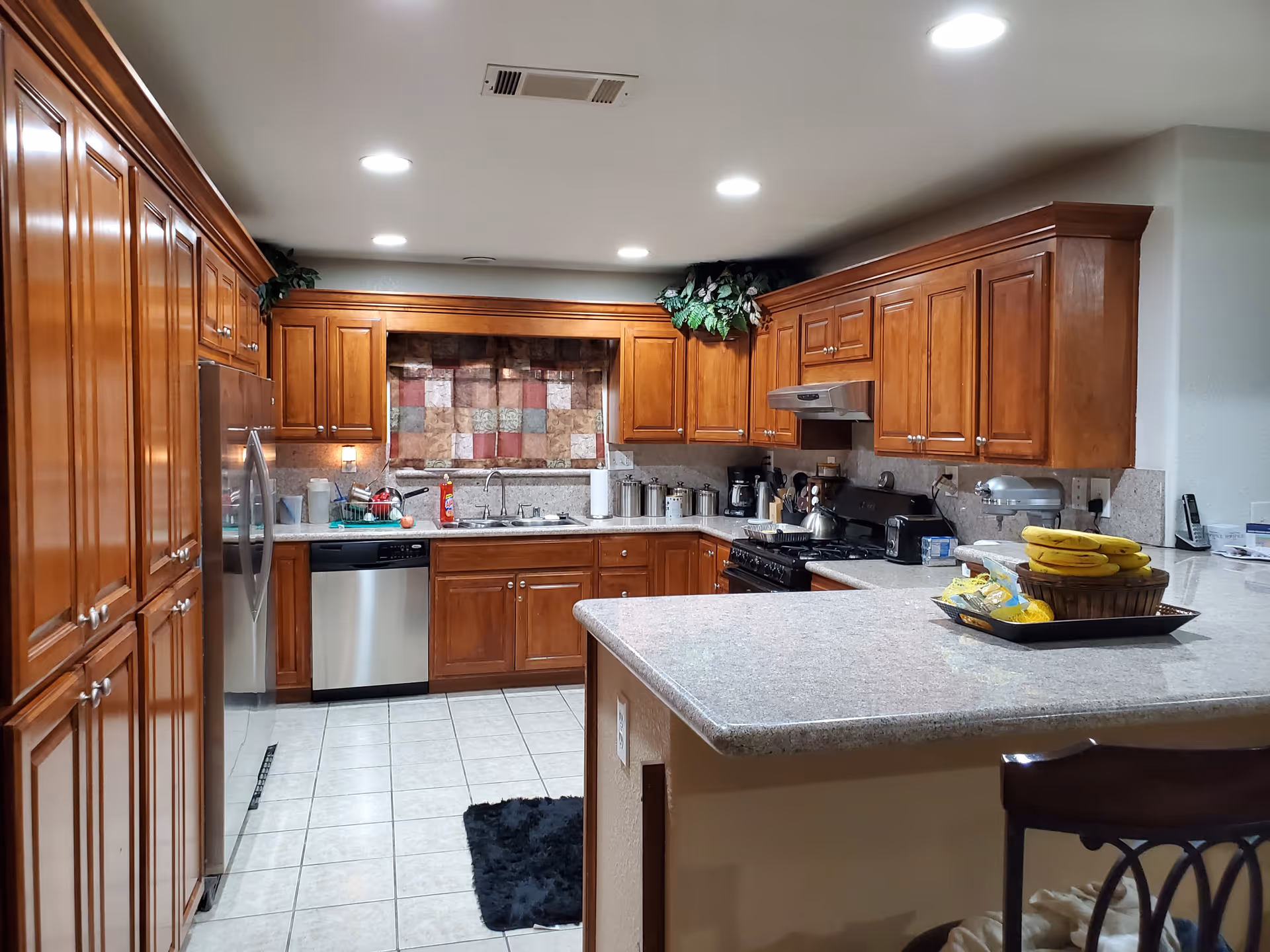 A spacious kitchen with wooden cabinets, stainless steel appliances including a refrigerator, dishwasher, and stove. The countertop is light-colored with a basket of bananas and other snacks. There is a window with a patchwork curtain above the sink, and the floor is tiled. The kitchen is well-lit with recessed ceiling lights.