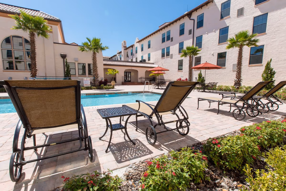 Outdoor pool area at Lake Gibson Village with several lounge chairs and small tables on a paved deck. Palm trees and flowering plants surround the pool, and a multi-story building with many windows is in the background under a clear blue sky.