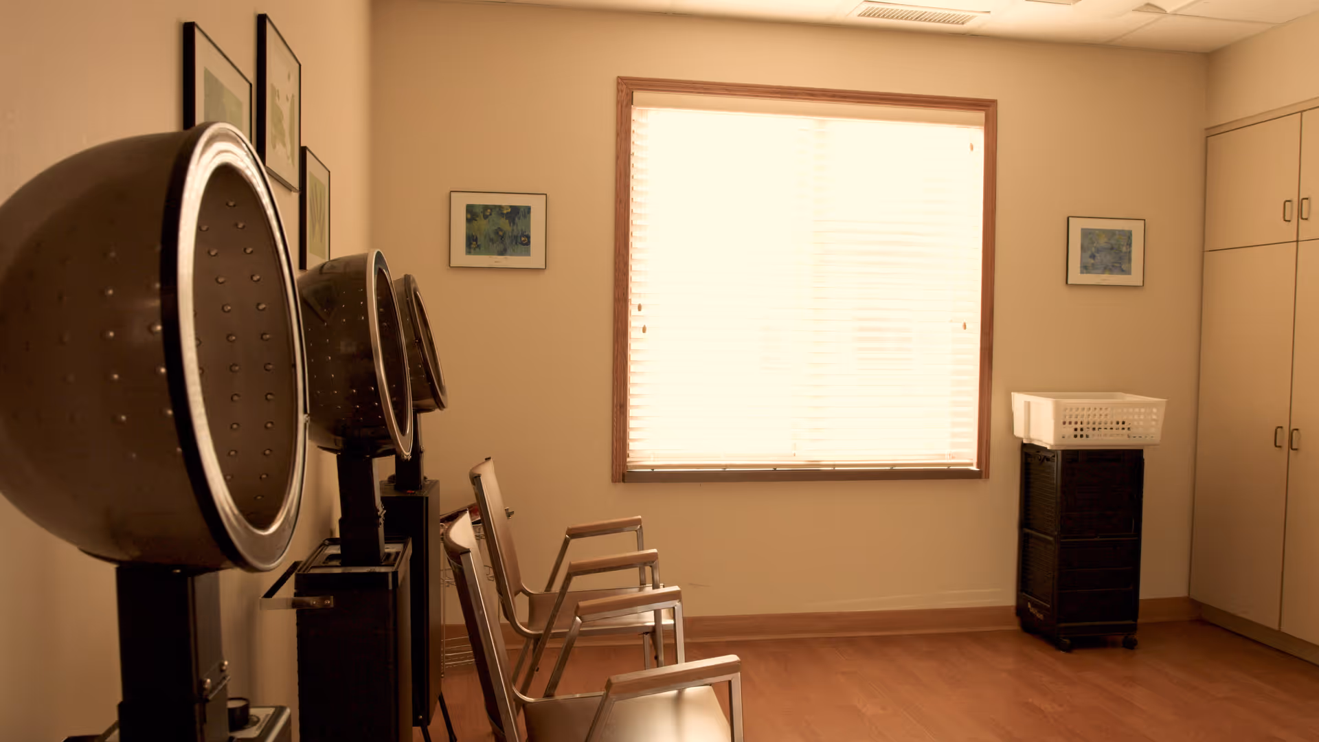Interior room with three vintage hair dryers on stands lined up against the left wall, three metal chairs positioned in front of them, a window with blinds letting in light, framed pictures on the walls, and a cabinet with a laundry basket on top on the right side.