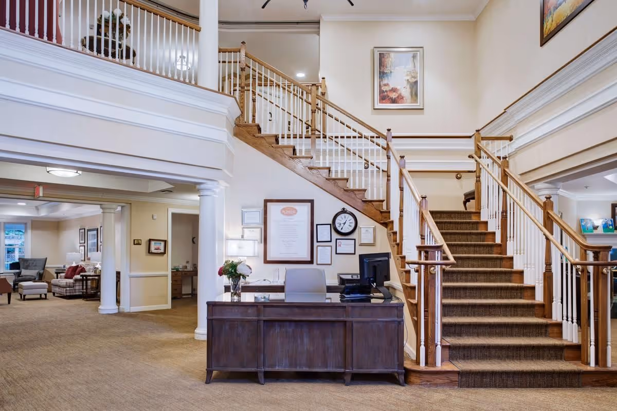 Interior view of a senior living facility lobby with a wooden reception desk in front of a staircase with wooden handrails and white balusters. The area has beige walls, carpeted floors, and framed artwork on the walls. To the left, there is a seating area with armchairs and a sofa, and white columns separate the spaces.