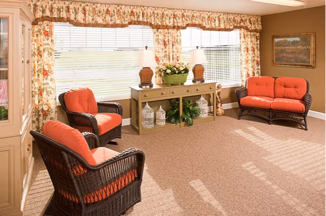A cozy living room area with three wicker chairs and a loveseat, all with orange cushions. There is a beige console table against the wall with two lamps, decorative birdcages, and a green plant. Large windows with floral curtains let in natural light, and a framed painting hangs on the wall.