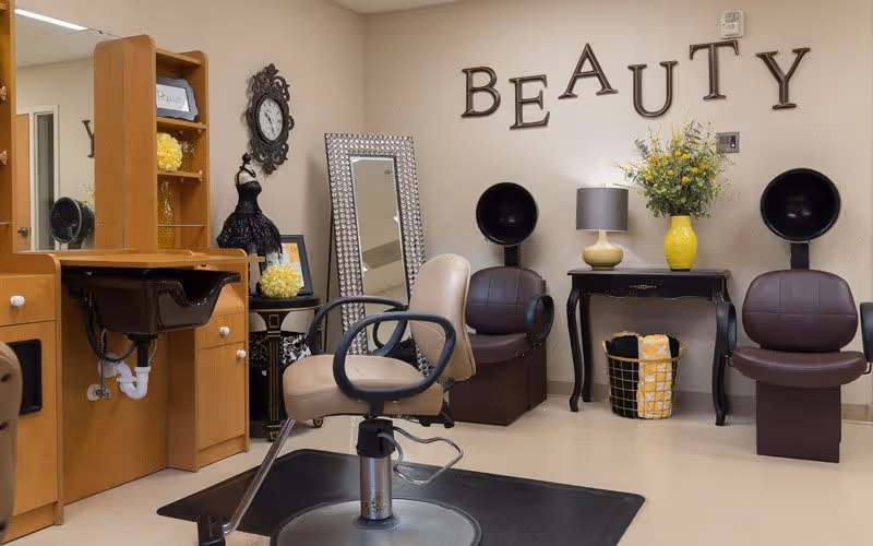 Interior view of a beauty salon area in a senior living facility with a styling chair in front of a wooden vanity with a mirror, two brown hair dryer chairs, a black table with a yellow vase of flowers and a lamp, a large standing mirror, and the word 'BEAUTY' displayed on the wall.