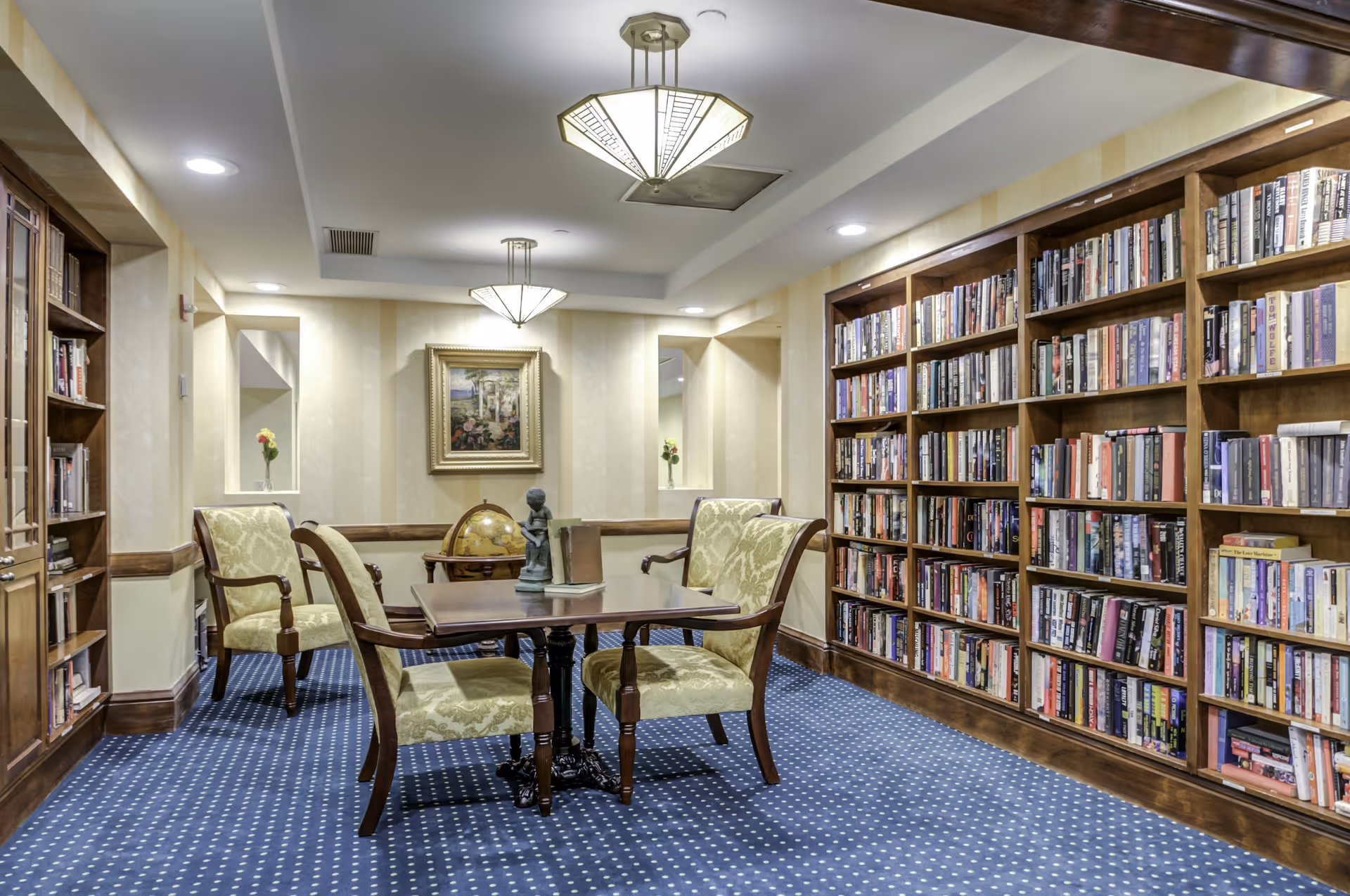 A cozy library room with wooden bookshelves filled with books along the right wall and a wooden cabinet with glass doors on the left. In the center, there is a wooden table with four upholstered chairs in a light green patterned fabric. On the table, there are a few books and a small statue. The room has a blue carpet with white polka dots, beige striped wallpaper, two modern ceiling lights, and a framed painting on the back wall above a globe stand.