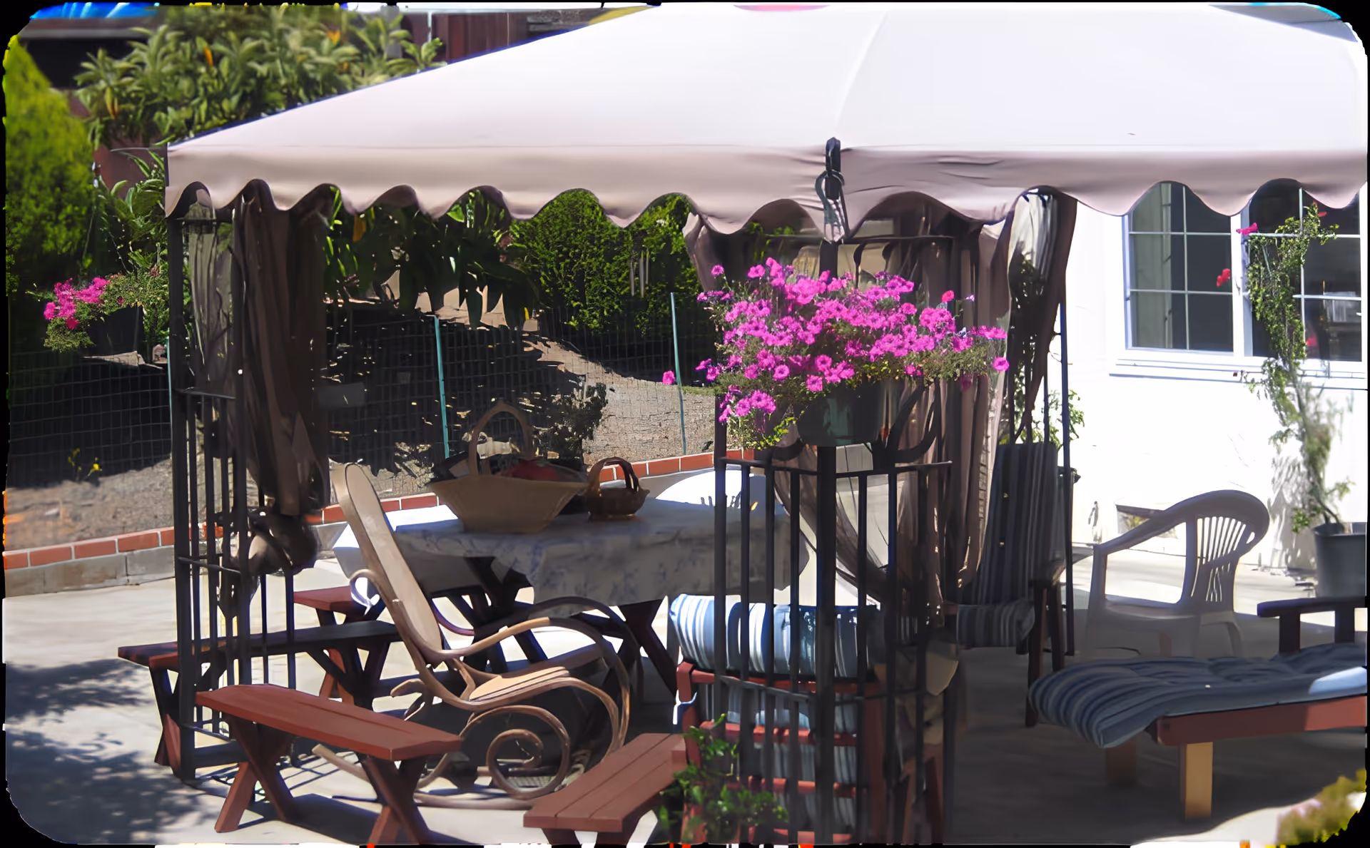 Outdoor shaded patio with a canopy, table and chairs, lounge seating and hanging pink flowers.