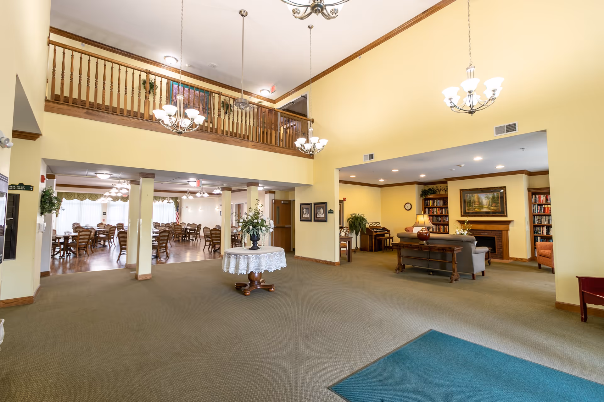 Spacious interior of a senior living facility with a large open area featuring a round table with a floral centerpiece in the middle. To the left, there is a dining area with multiple wooden tables and chairs. To the right, there is a cozy sitting area with sofas, a lamp, bookshelves, and a fireplace. The walls are painted light yellow, and the ceiling is high with hanging chandeliers. A wooden railing balcony is visible above the dining area.