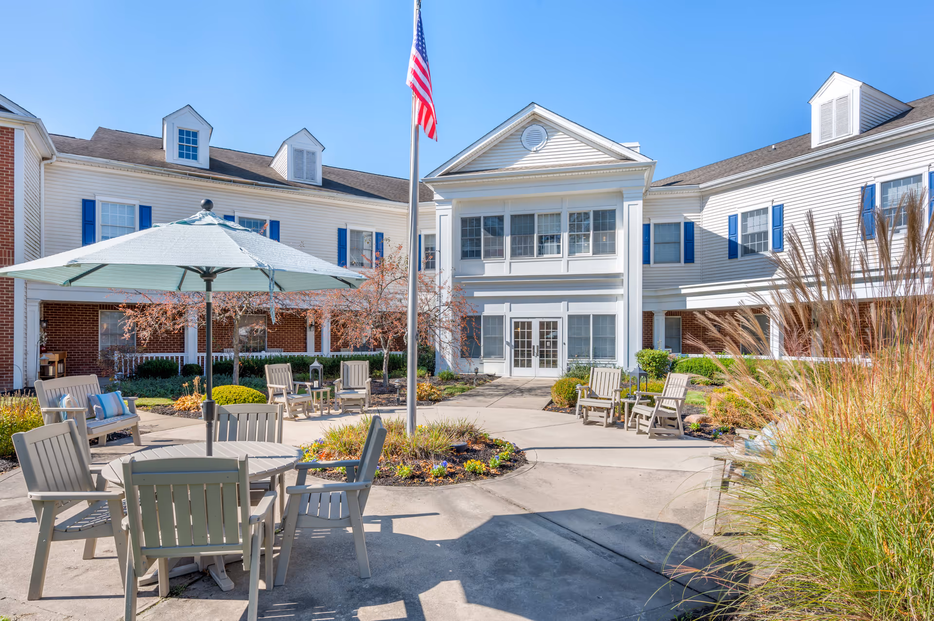 Outdoor courtyard area of a senior living facility with multiple seating arrangements including tables with umbrellas and chairs, surrounded by landscaped plants and bushes. The building in the background is white with blue shutters and has multiple windows. An American flag is prominently displayed in the center of the courtyard.
