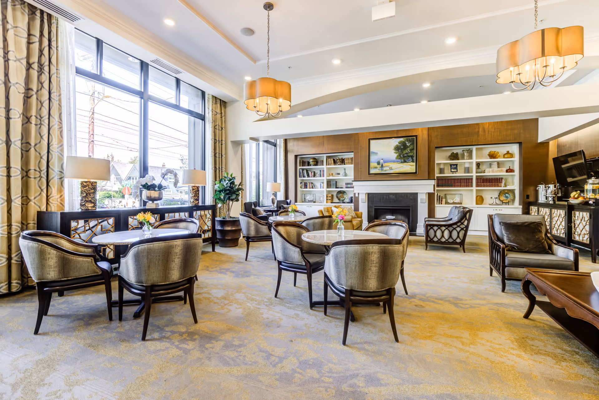 A spacious and well-lit common area in a senior living facility featuring multiple round tables with upholstered chairs, large windows with patterned curtains, two hanging chandeliers, a fireplace with built-in bookshelves on either side, and comfortable armchairs arranged around the room. There are decorative lamps, plants, and a painting above the fireplace.
