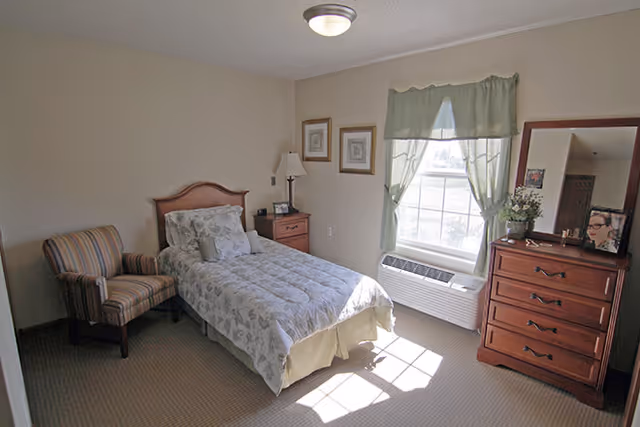 Sunlit furnished bedroom with a single bed, striped armchair, wooden dresser, and a window with green curtains.