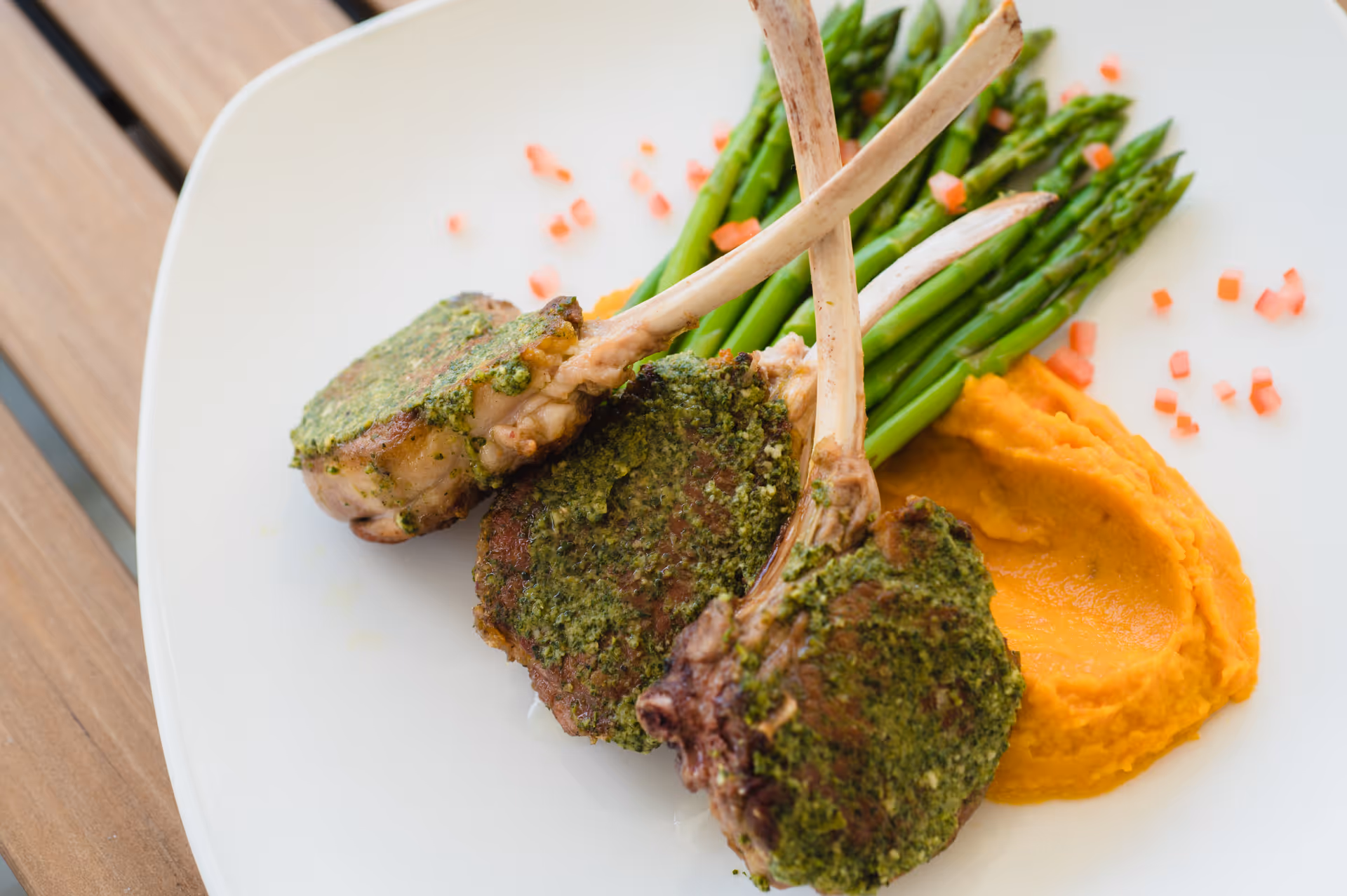 A white plate with three herb-crusted lamb chops, a serving of mashed sweet potatoes, and a bundle of steamed asparagus, garnished with small diced red peppers.