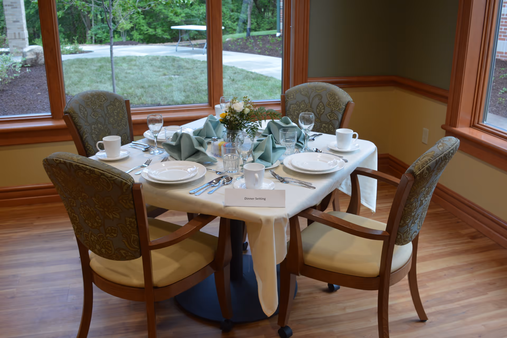 A dining table set for four with white plates, cups, silverware, glasses, and folded green napkins. A small floral centerpiece is in the middle of the table. The table is in a room with large windows showing a green outdoor area.