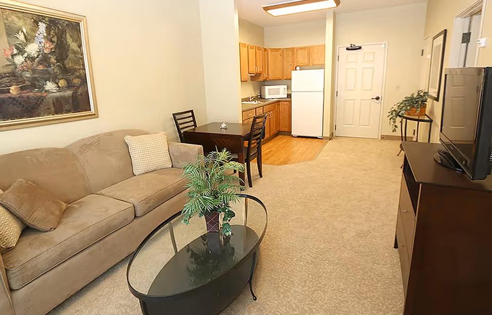 Cozy senior apartment living area with sofa, coffee table, plant, TV, and kitchenette.