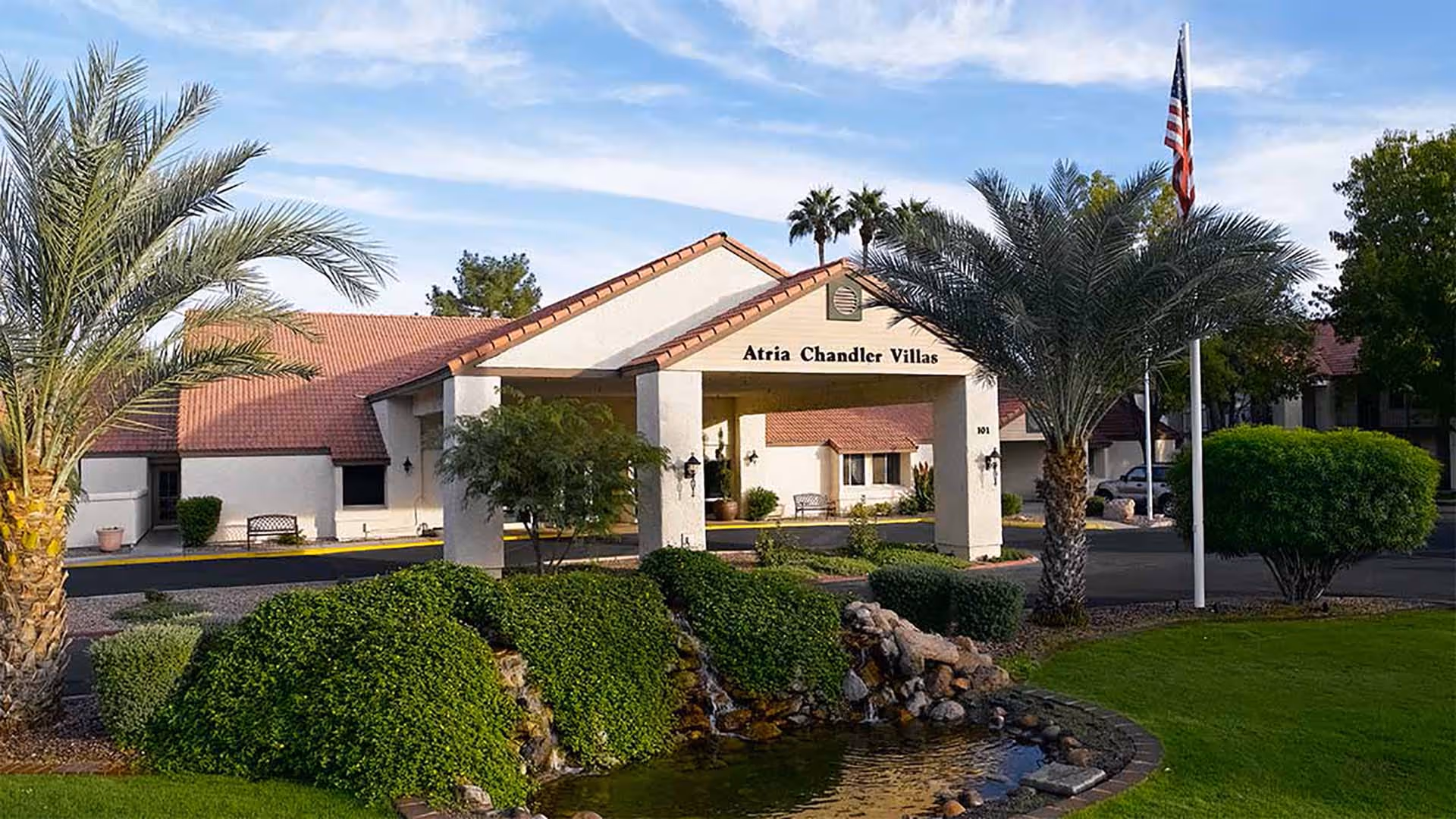 The landscaped front entrance of Atria Chandler Villas showing a covered porte-cochère, palm trees, and a small pond.