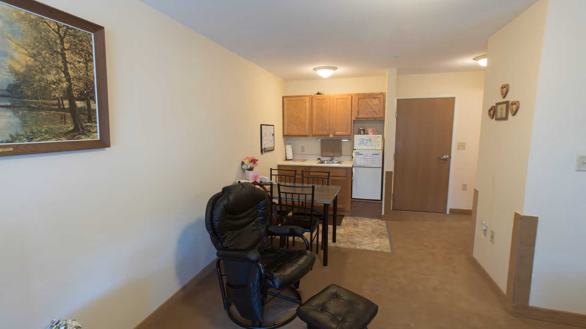 Interior view of a small living space featuring a black cushioned chair with matching footrest, a dining table with four chairs, and a kitchenette with wooden cabinets, a sink, a microwave, and a refrigerator. The walls are light-colored with minimal decorations including a framed landscape painting and small heart-shaped wall hangings. The floor is carpeted except for the kitchenette area which has tiled flooring.